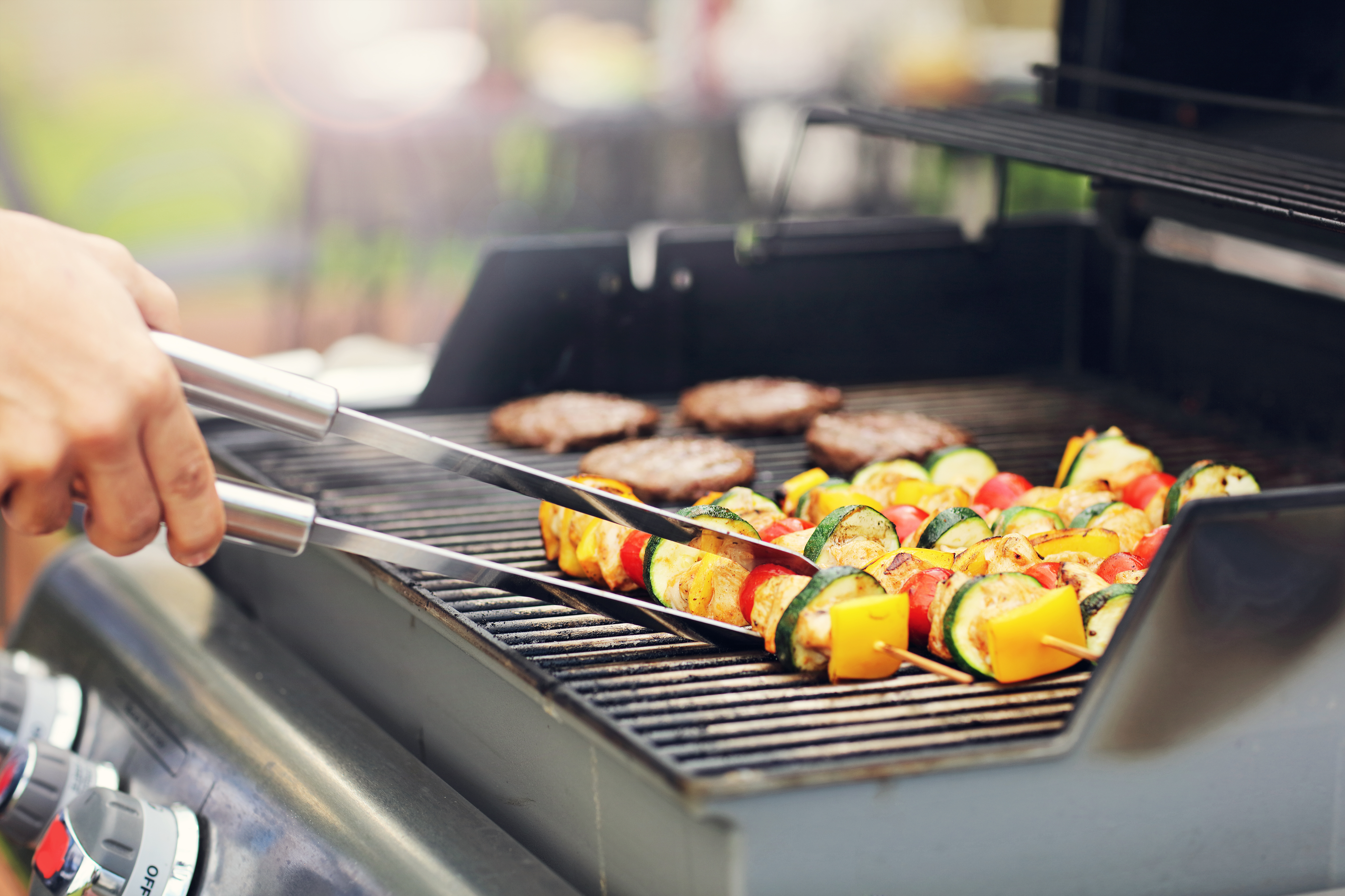 Ein Griller mit Gemüsespießen und Burger-Patties im Hintergrund