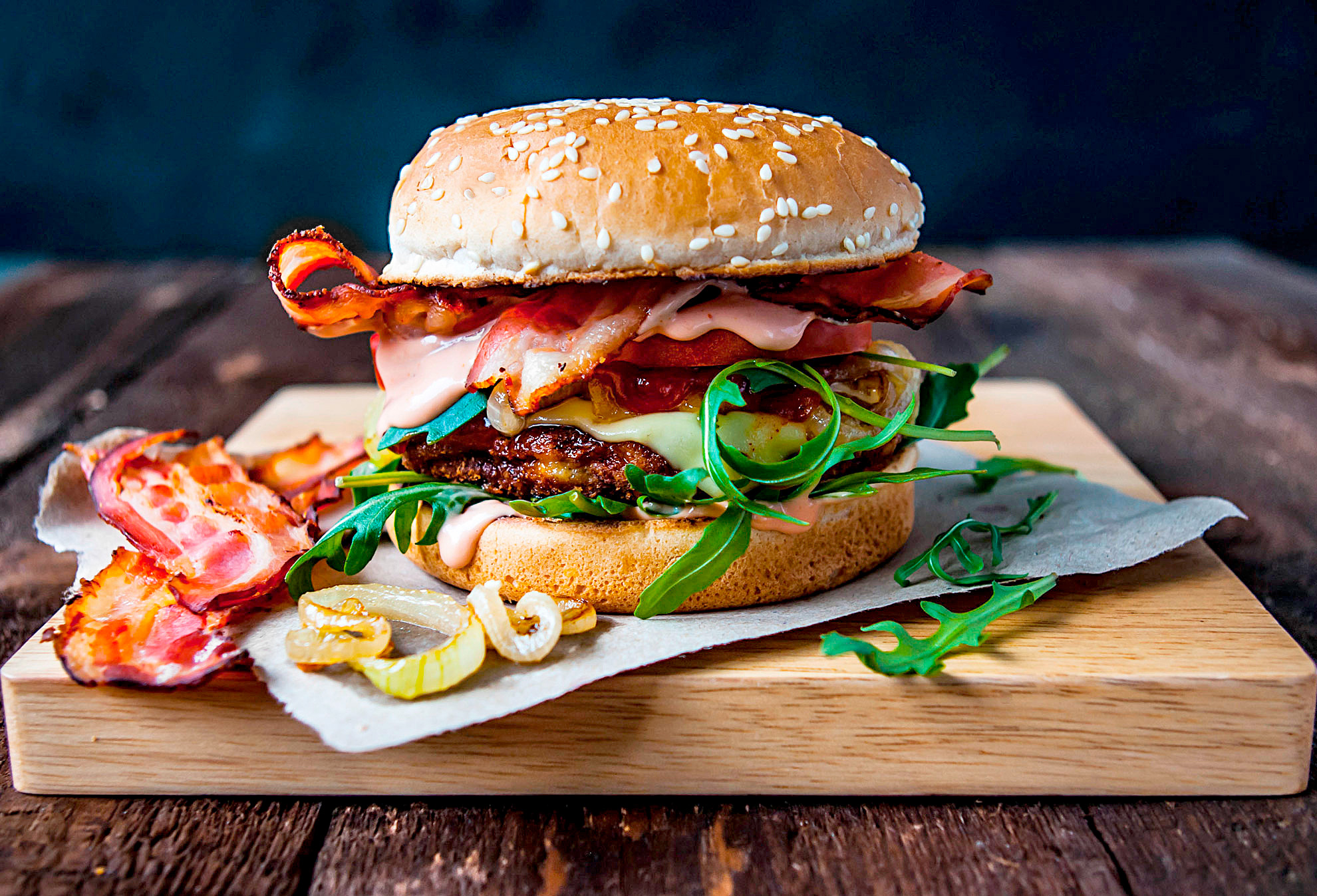 Ein Burger mit Speck, Salat, Zwiebel, Käse und Sauce liegt auf einem Holzbrett