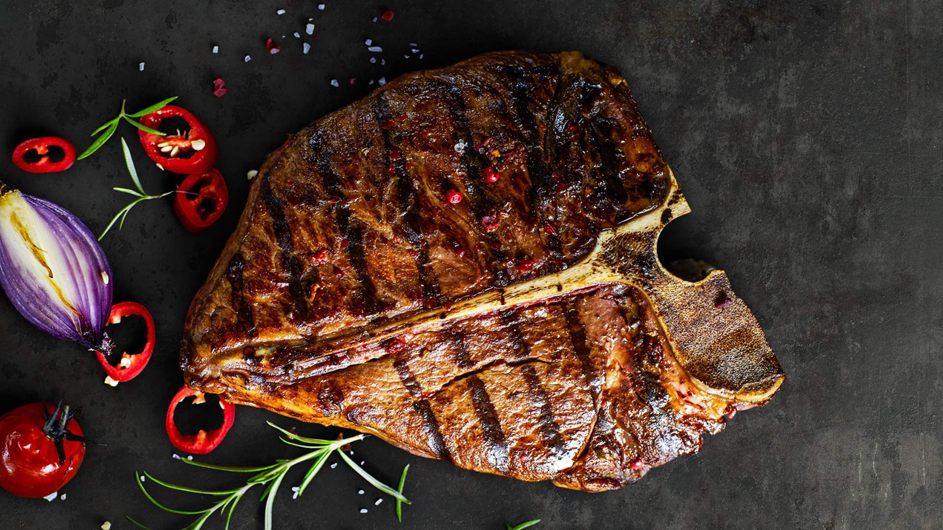 Gegrilltes Steak liegt auf einer dunkler Platte, rundherum sind Kräuter, Zwiebel und Salz verstreut