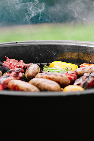 Ein runder Kohlegrill mit Würstel und Paprikastücken steht in einem Garten